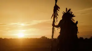 The silhouette of a native american man dressed in regalia holding a staff sitting on a horse as the sun sets in the background