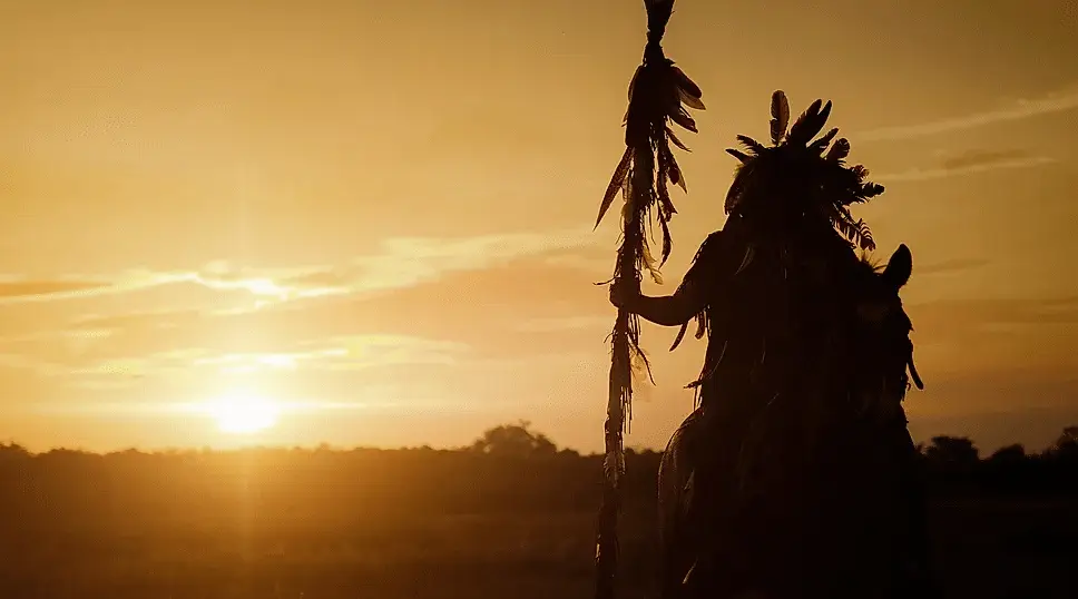 The silhouette of a native american man dressed in regalia holding a staff sitting on a horse as the sun sets in the background