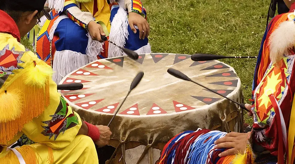 Group of Native American drummers in traditional regalia playing a large, decorated drum at a cultural gathering.