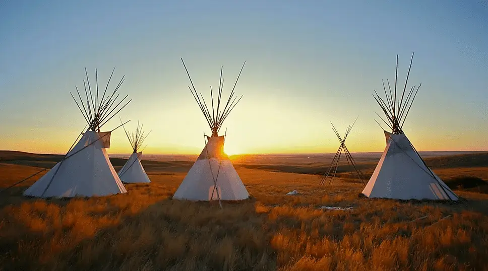 Traditional Native American tipis in an open grassland at sunset, with the sunrise casting a golden glow across the landscape.