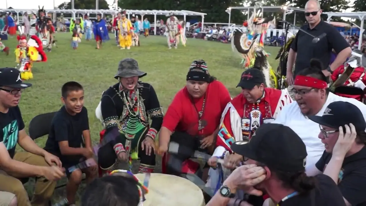 Group of Native American men and boys participating in a traditional drumming circle at a powwow, with dancers and spectators in the background.