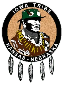 Logo of the Iowa Tribe of Kansas and Nebraska featuring a depiction of Chief White Cloud wearing traditional regalia, surrounded by a circular black band with the text 'Iowa Tribe' at the top and 'Kansas - Nebraska' at the bottom. The logo is adorned with five feathers hanging from the bottom of the circle.