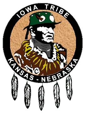 Logo of the Iowa Tribe of Kansas and Nebraska featuring a depiction of Chief White Cloud wearing traditional regalia, surrounded by a circular black band with the text 'Iowa Tribe' at the top and 'Kansas - Nebraska' at the bottom. The logo is adorned with five feathers hanging from the bottom of the circle.