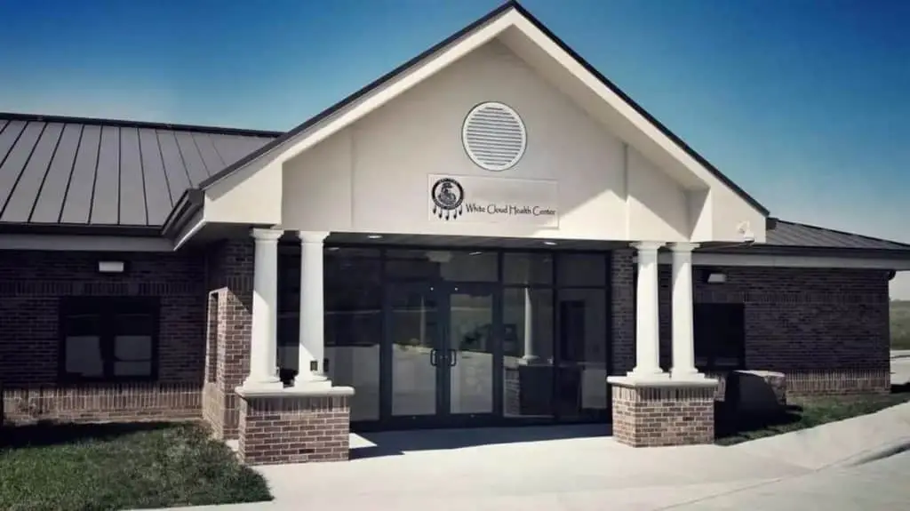 The front entrance of White Cloud Health Center which is a brick building with glass doors, 4 columns at the entrance, a brown roof, under an open blue sky.