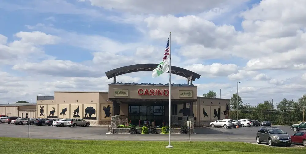 A large casino building with animal silhouettes on the exterior walls, located under a partly cloudy sky. An American flag and the Iowa Tribe of Kansas and Nebraska flag stand tall in front of the building, surrounded by a parking lot with several cars.