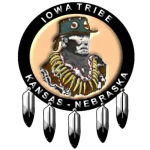 Logo of the Iowa Tribe of Kansas and Nebraska featuring a depiction of Chief White Cloud wearing traditional regalia, surrounded by a circular black band with the text 'Iowa Tribe' at the top and 'Kansas - Nebraska' at the bottom. The logo is adorned with five feathers hanging from the bottom of the circle.