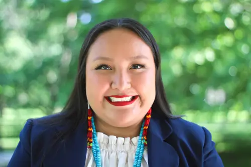 A professional headshot of a woman with long dark hair, smiling brightly and wearing traditional beaded jewelry with a navy blazer, set against a blurred, vibrant green outdoor background.