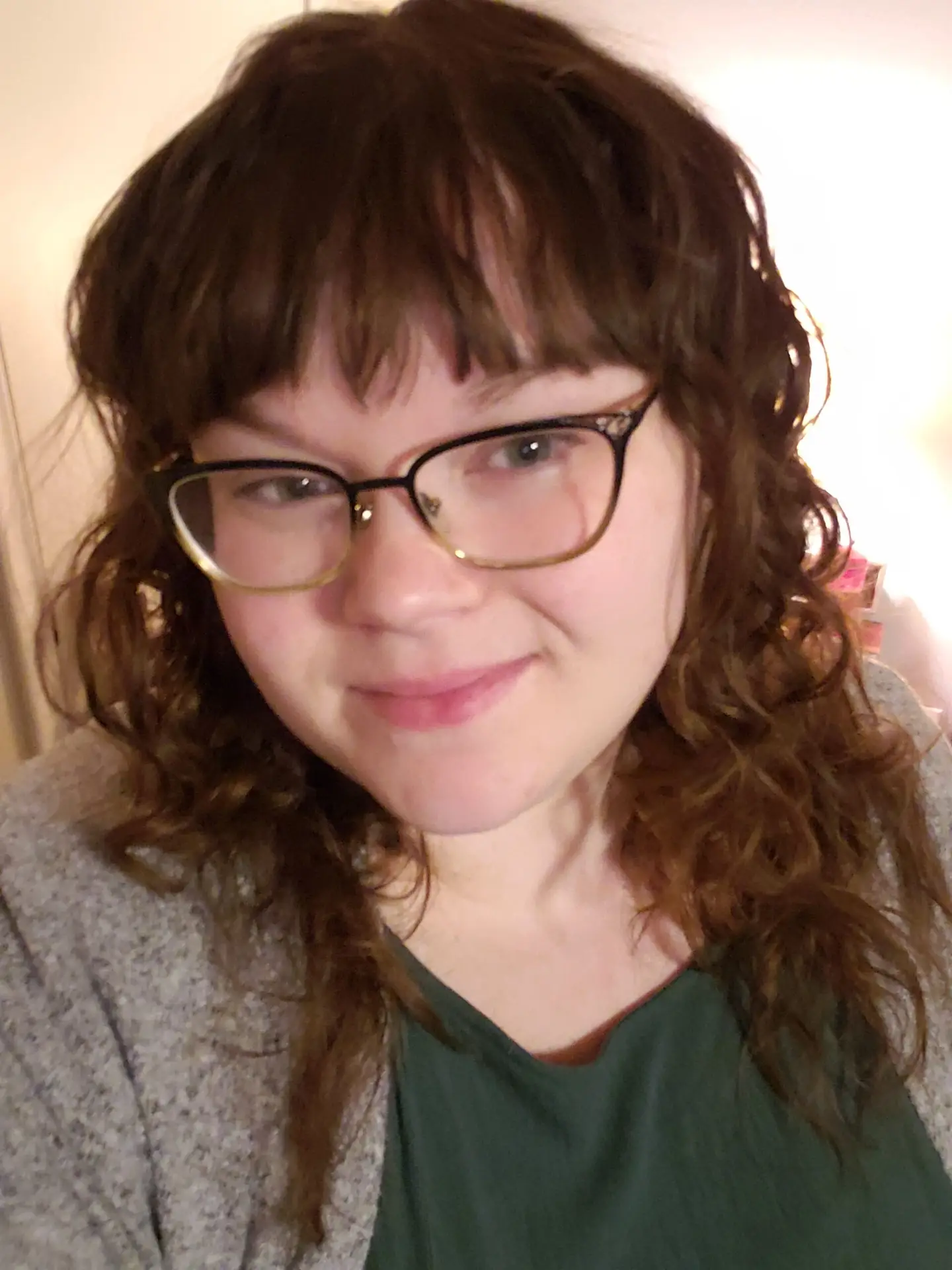 A casual headshot of a woman with curly brown hair and glasses, smiling softly, wearing a green top and gray sweater in a warmly lit indoor setting.