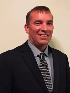 Vice-Chairman Brandon Roberts in a dark suit and patterned tie, smiling confidently, standing against a neutral background.