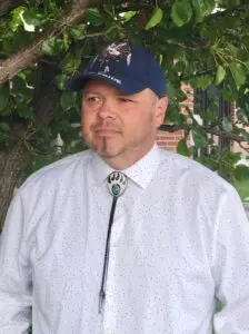 Chairman Timothy Rhodd wearing a blue baseball cap, white shirt, and a bolo tie stands outdoors in front of a tree, looking to the side with a serious expression.