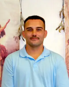 Secretary Tony Fee in a light blue polo shirt with a mustache, standing in front of a backdrop featuring Native American cultural imagery and hanging feathers.