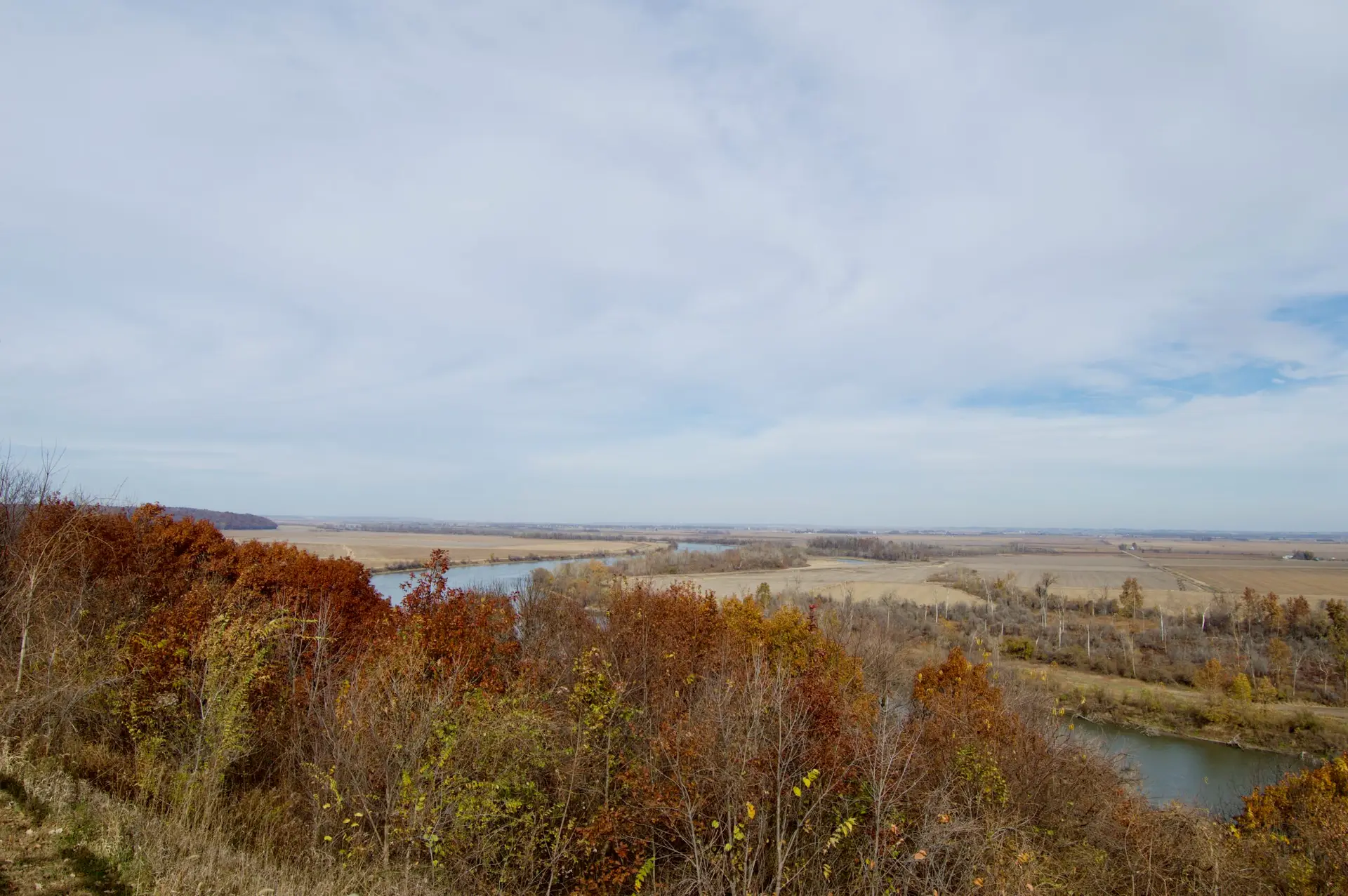 A scenic view of the Missouri River winding through a flat, open landscape with autumn trees in the foreground and vast fields stretching into the distance under a partly cloudy sky.