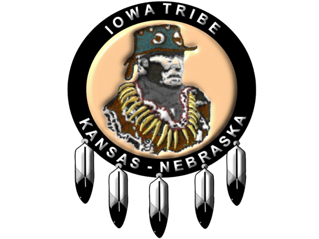 Logo of the Iowa Tribe of Kansas and Nebraska featuring a depiction of Chief White Cloud wearing traditional regalia, surrounded by a circular black band with the text 'Iowa Tribe' at the top and 'Kansas - Nebraska' at the bottom. The logo is adorned with five feathers hanging from the bottom of the circle.