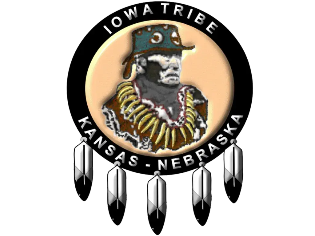 Logo of the Iowa Tribe of Kansas and Nebraska featuring a depiction of Chief White Cloud wearing traditional regalia, surrounded by a circular black band with the text 'Iowa Tribe' at the top and 'Kansas - Nebraska' at the bottom. The logo is adorned with five feathers hanging from the bottom of the circle.