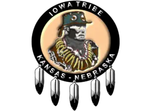 Logo of the Iowa Tribe of Kansas and Nebraska featuring a depiction of Chief White Cloud wearing traditional regalia, surrounded by a circular black band with the text 'Iowa Tribe' at the top and 'Kansas - Nebraska' at the bottom. The logo is adorned with five feathers hanging from the bottom of the circle.