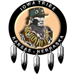 Logo of the Iowa Tribe of Kansas and Nebraska featuring a depiction of Chief White Cloud wearing traditional regalia, surrounded by a circular black band with the text 'Iowa Tribe' at the top and 'Kansas - Nebraska' at the bottom. The logo is adorned with five feathers hanging from the bottom of the circle.