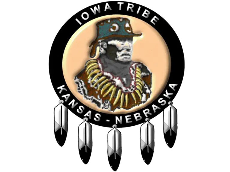 Logo of the Iowa Tribe of Kansas and Nebraska featuring a depiction of Chief White Cloud wearing traditional regalia, surrounded by a circular black band with the text 'Iowa Tribe' at the top and 'Kansas - Nebraska' at the bottom. The logo is adorned with five feathers hanging from the bottom of the circle.