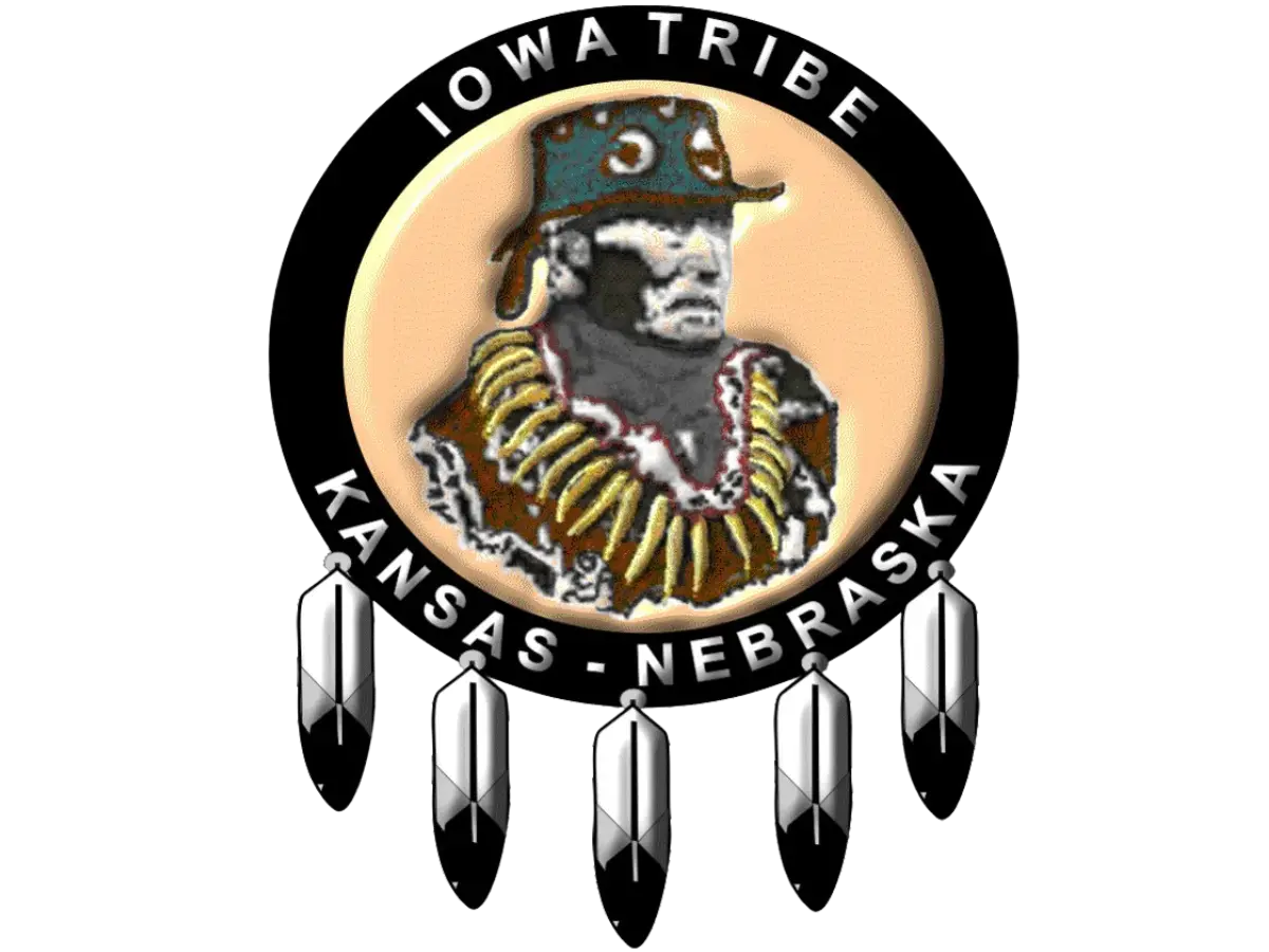 Logo of the Iowa Tribe of Kansas and Nebraska featuring a depiction of Chief White Cloud wearing traditional regalia, surrounded by a circular black band with the text 'Iowa Tribe' at the top and 'Kansas - Nebraska' at the bottom. The logo is adorned with five feathers hanging from the bottom of the circle.