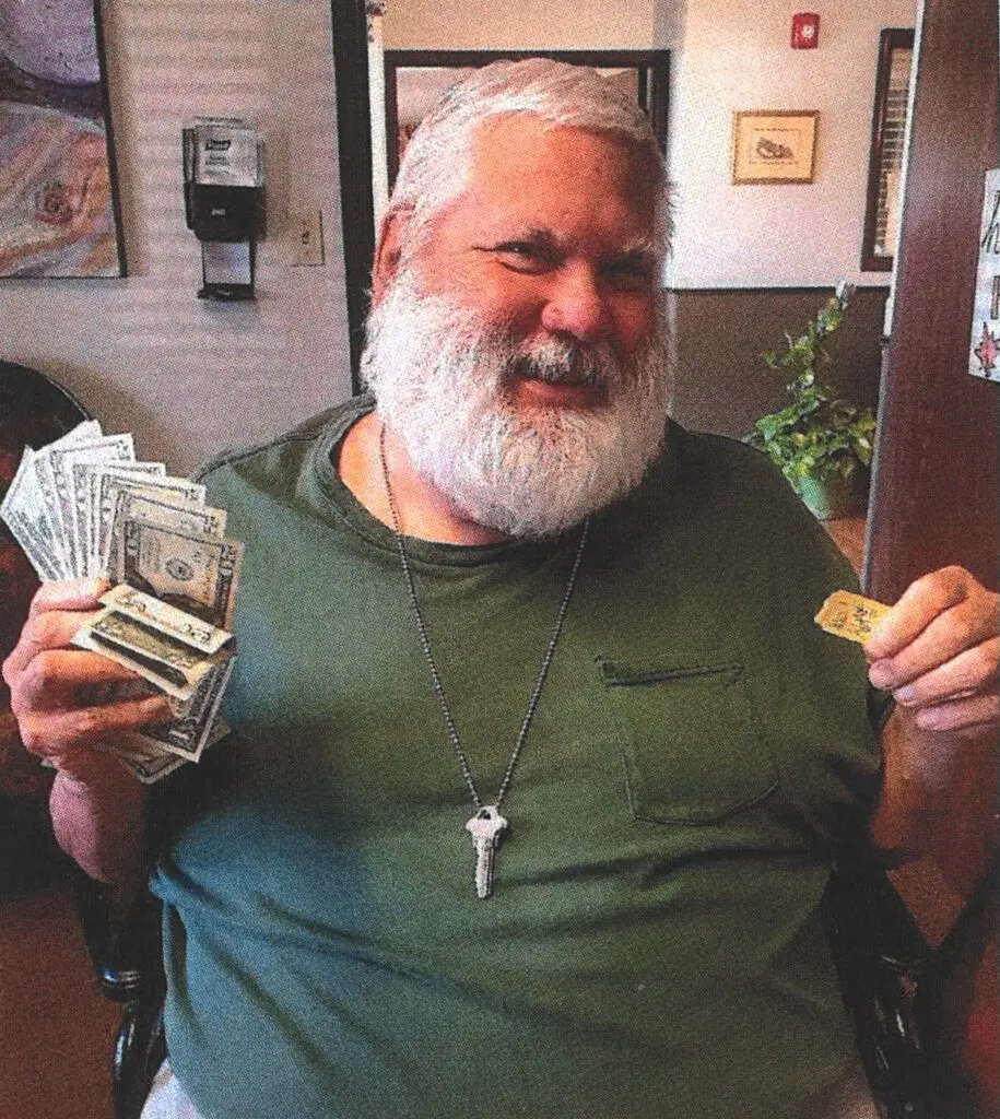 The image is of Bill Jensen with a white beard, wearing a green pocketed t-shirt, with a key around his neck, holding up a raffle ticket and cash. The background is an office setting.