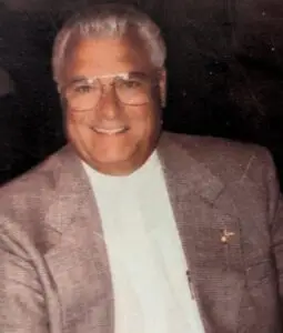 An image of William "Bill" Harvey Campbell smiling with a tweed jacket and white shirt on with a dark background.