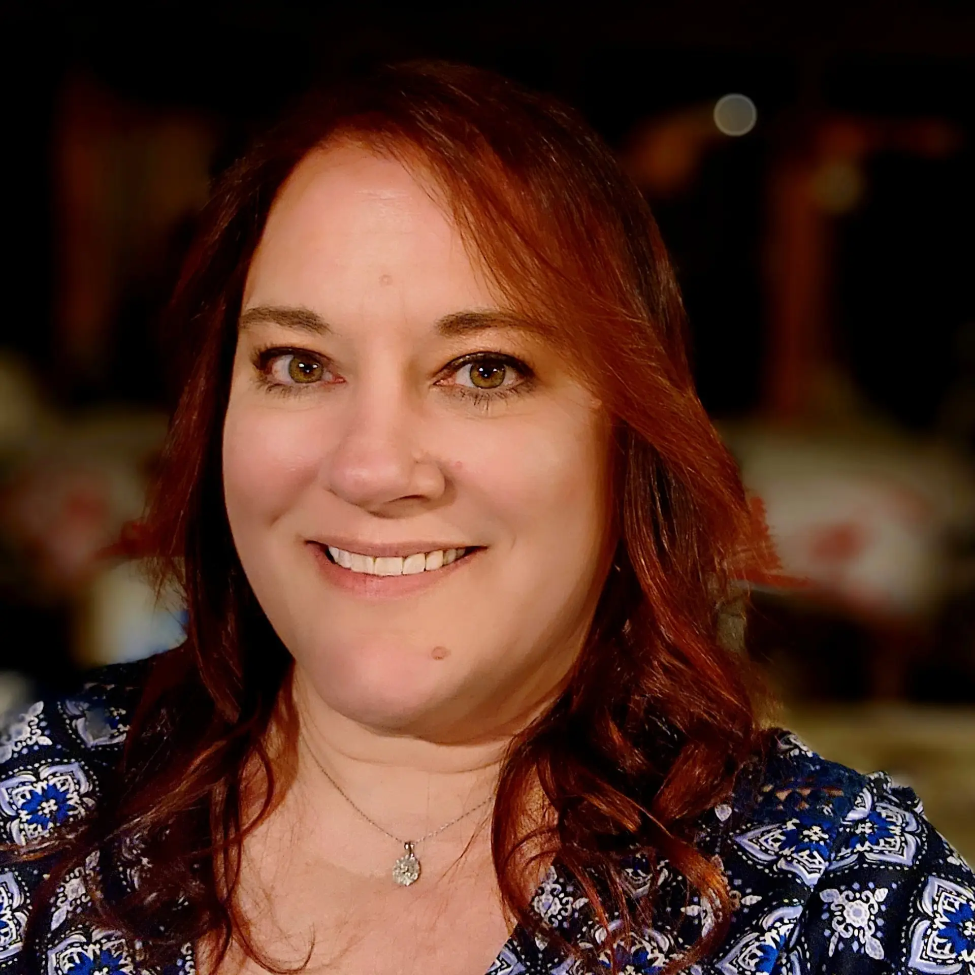 A smiling woman with shoulder-length auburn hair, wearing a blue patterned top and a silver necklace, poses in a warm, softly lit environment.