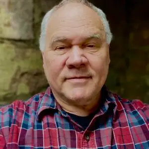 A middle-aged man with short, light-colored hair is wearing a red and blue plaid shirt. He is standing outdoors in front of a stone wall, looking directly at the camera with a calm expression.