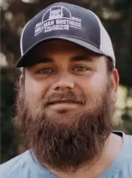 Treasurer Robert Hullman with a full beard, wearing a baseball cap with the logo "Hillman Brothers Farm" on it, smiling softly at the camera.