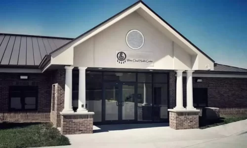 The front entrance of White Cloud Health Center which is a brick building with glass doors, 4 columns at the entrance, a brown roof, under an open blue sky.