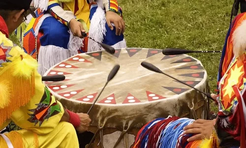 Group of Native American drummers in traditional regalia playing a large, decorated drum at a cultural gathering.