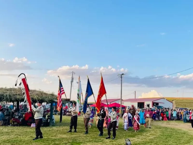 Native American powwow with a color guard procession carrying flags, surrounded by attendees in traditional regalia, musicians, and spectators in an open field under a clear sky.