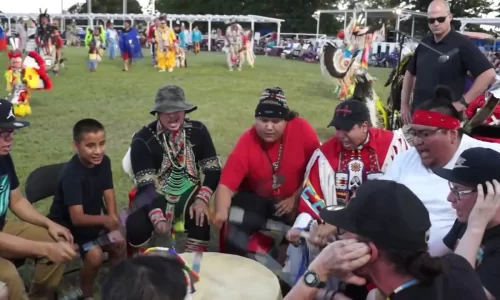 Group of Native American men and boys participating in a traditional drumming circle at a powwow, with dancers and spectators in the background.